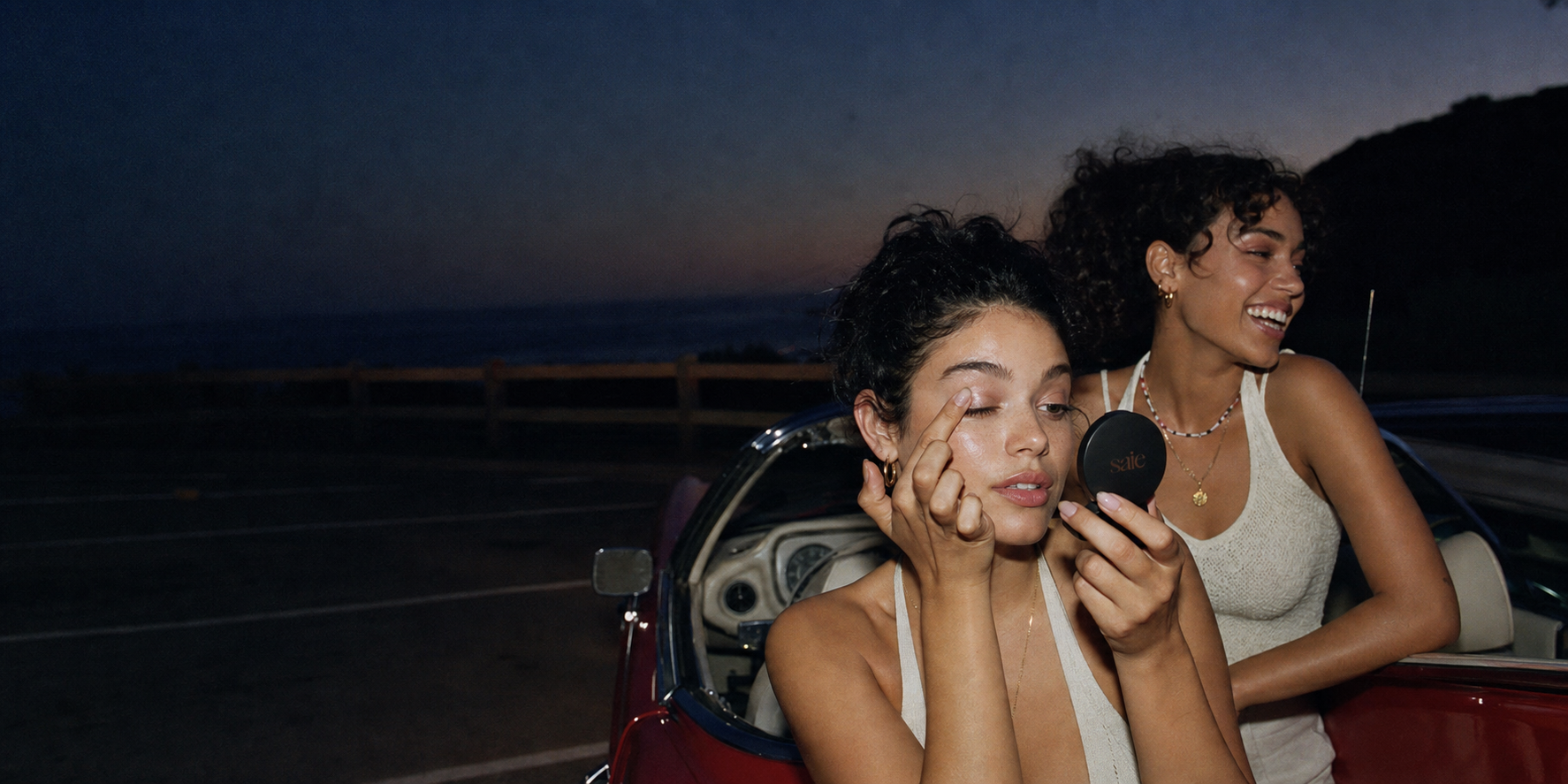 Friends applying eyeshadow beside a convertible after sunset